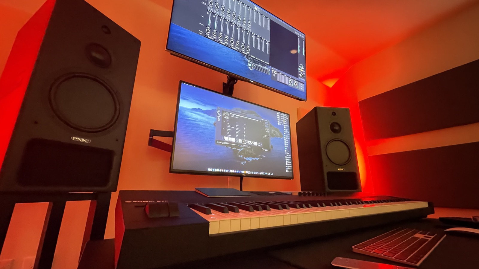 Recording studio with monitor speakers, keyboards, and two displays under warm red lighting.