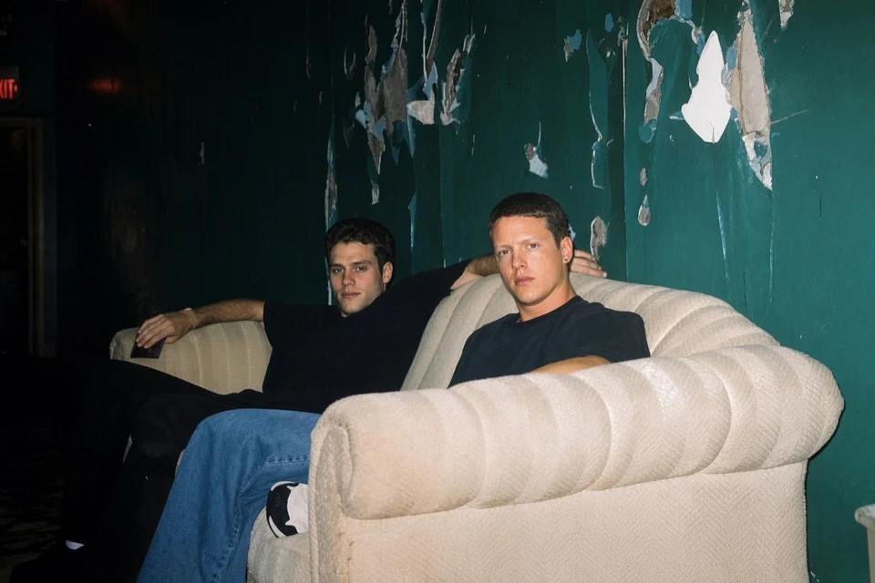 Two people seated on a cream sofa against a weathered teal wall